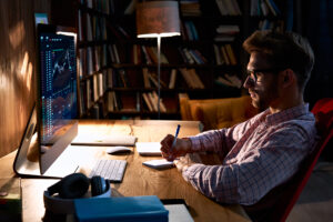 A man with glasses and a beard sits at a wooden desk in a dimly lit room, writing in a notebook . He is looking at a computer monitor displaying financial charts. A lamp illuminates the desk, and a bookshelf filled with books is visible in the background.