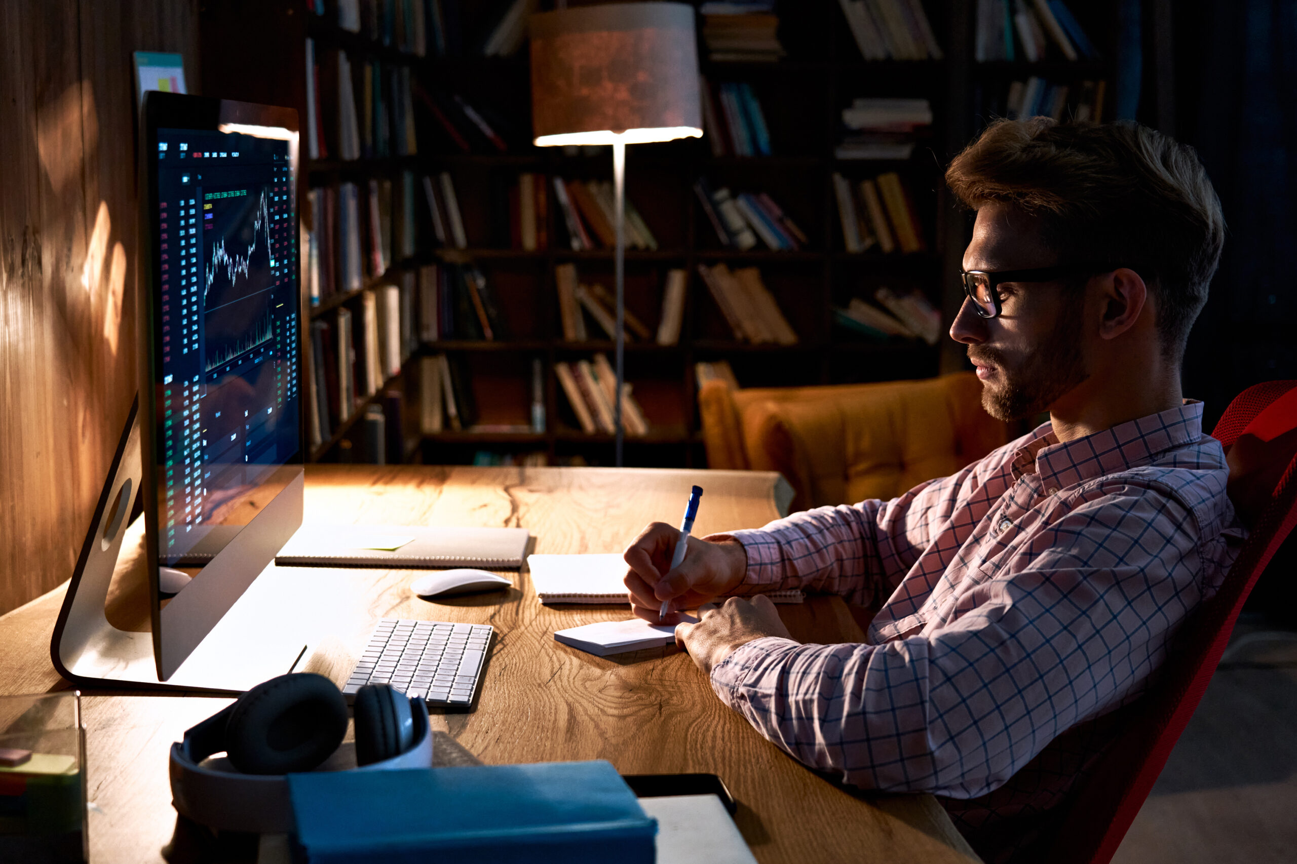 A man with glasses and a beard sits at a wooden desk in a dimly lit room, writing in a notebook . He is looking at a computer monitor displaying financial charts. A lamp illuminates the desk, and a bookshelf filled with books is visible in the background.